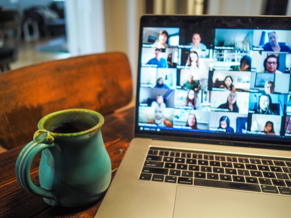 Laptop showing many people on a virtual group conference call with coffee cup next to it.